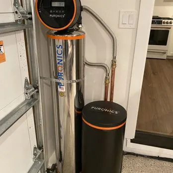 A water treatment system by Puronics, LLC installed in a residential setting, featuring a sleek metallic design with connected pipes and a control panel.